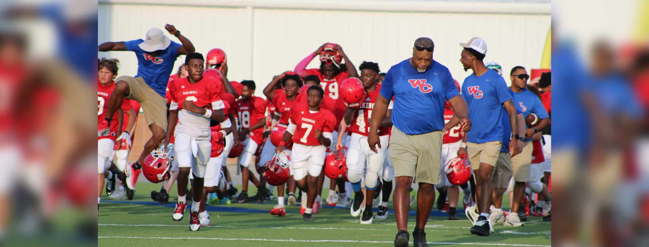 Warren Central Junior High's seventh-grade football team runs onto the field, wearing red jerseys and helmets, with coache…