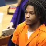 An 18-year-old in an orange jumpsuit sits in a courtroom, looking serious during sentencing. — 18 year old