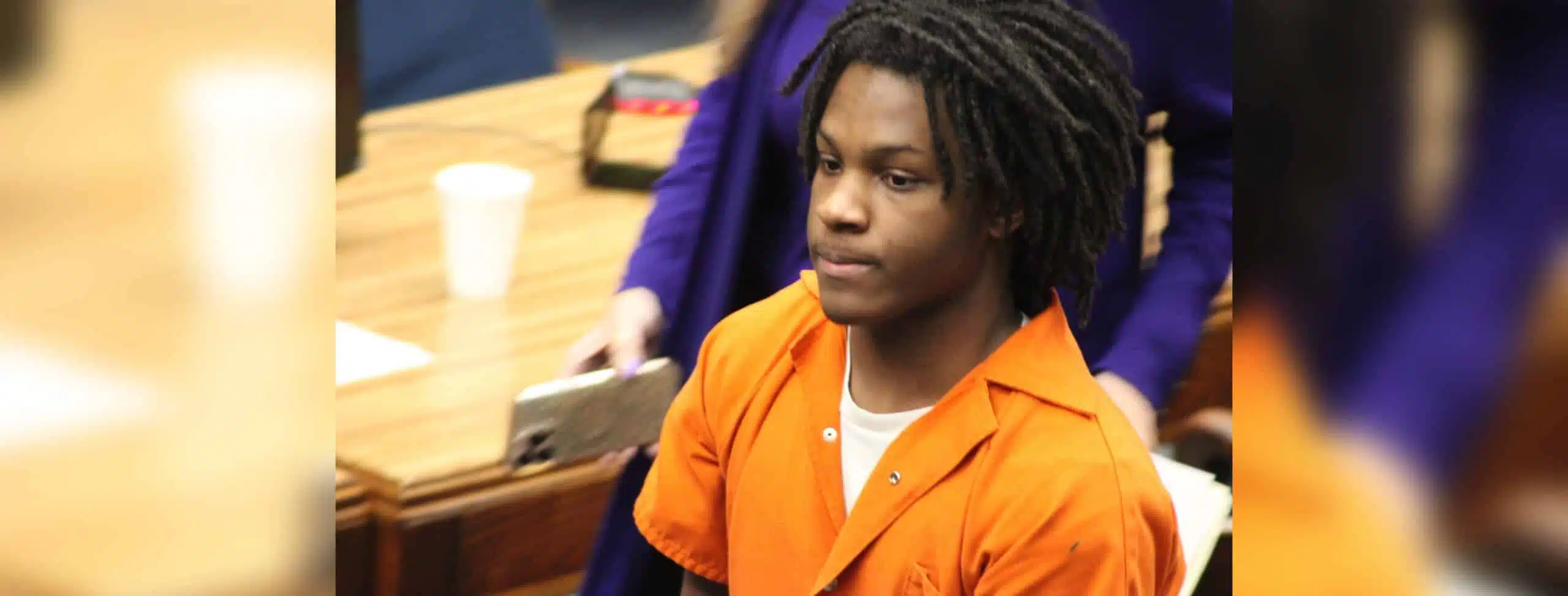 An 18-year-old in an orange jumpsuit sits in a courtroom, looking serious during sentencing. — 18 year old