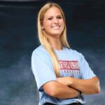 A confident volleyball player in a blue shirt stands with arms crossed, smiling against a dark background. — Sweeps canton