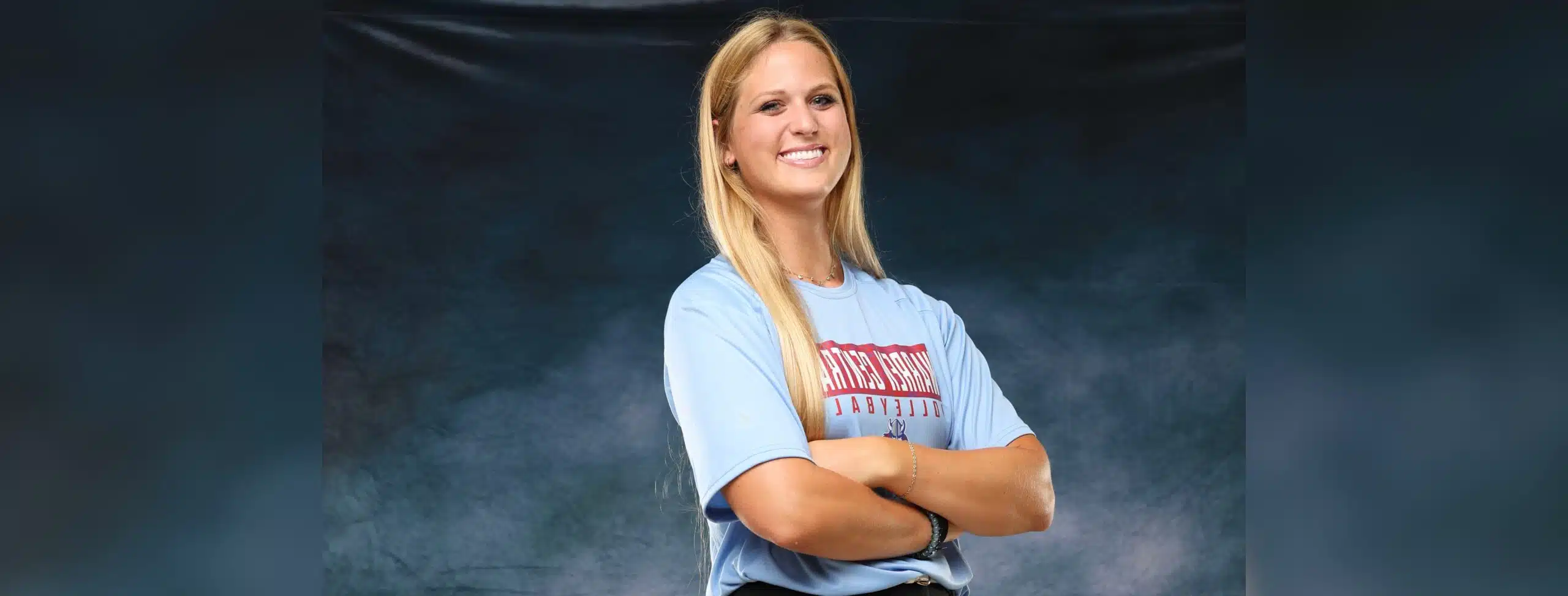 A confident volleyball player in a blue shirt stands with arms crossed, smiling against a dark background. — Sweeps canton
