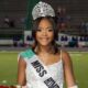 Lauren Gibbs wearing a crown and sash, smiling while holding flowers on a football field.