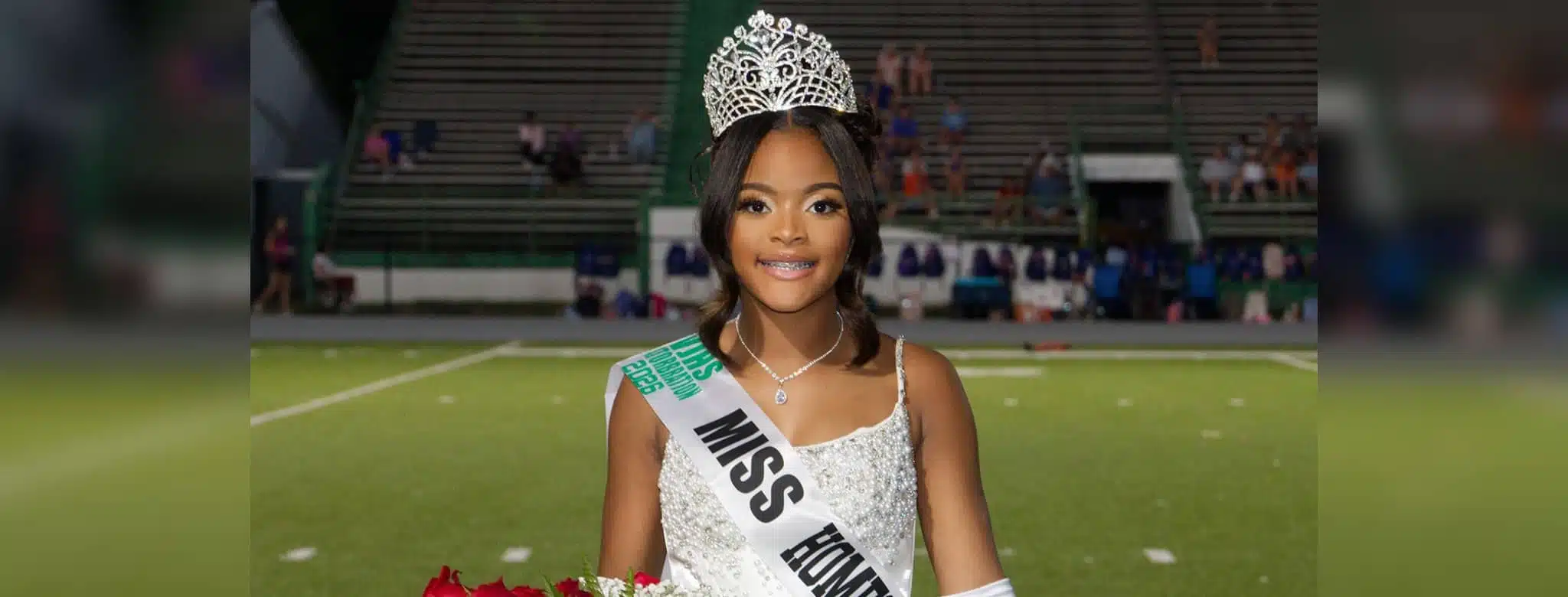 Lauren Gibbs wearing a crown and sash, smiling while holding flowers on a football field.