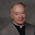 Father Robert Dore, a priest in a gray suit with a clerical collar, smiling softly at the camera.