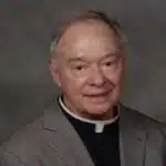 Father Robert Dore, a priest in a gray suit with a clerical collar, smiling softly at the camera.