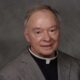 Father Robert Dore, a priest in a gray suit with a clerical collar, smiling softly at the camera.