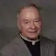 Father Robert Dore, a priest in a gray suit with a clerical collar, smiling softly at the camera.