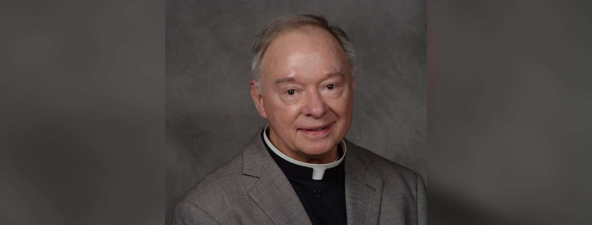 Father Robert Dore, a priest in a gray suit with a clerical collar, smiling softly at the camera.
