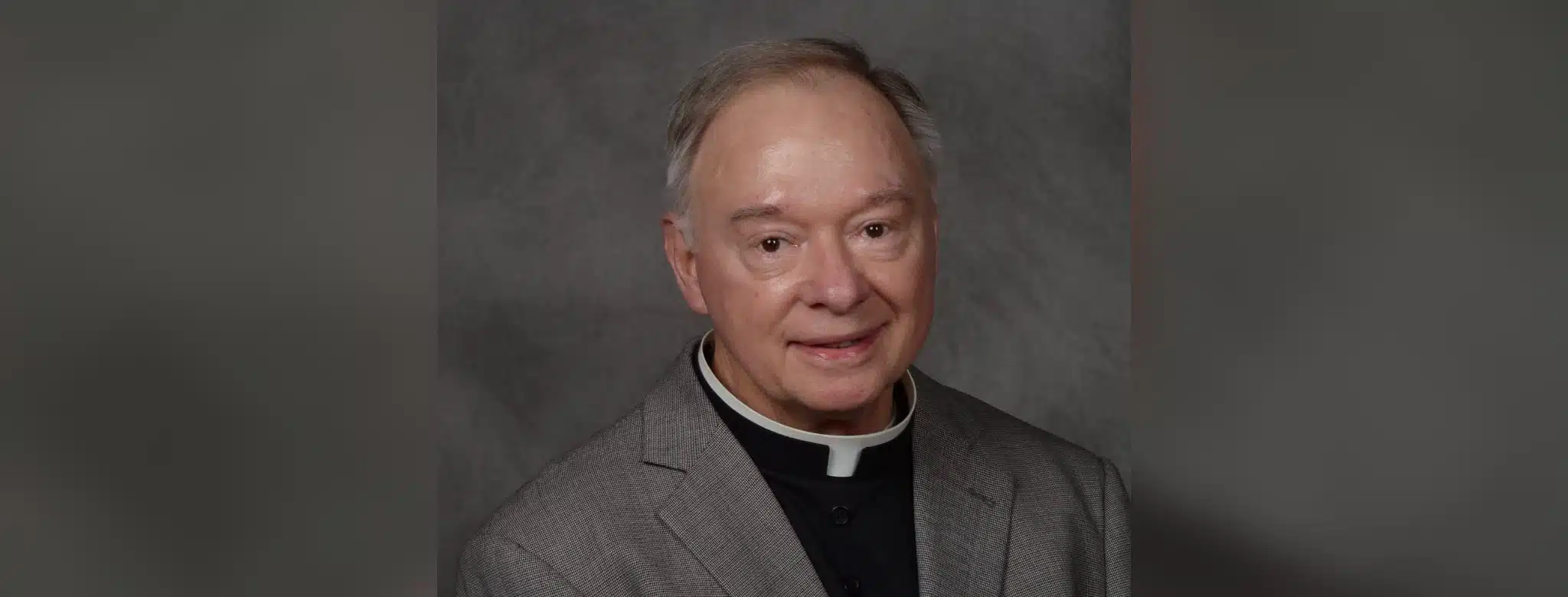 Father Robert Dore, a priest in a gray suit with a clerical collar, smiling softly at the camera.