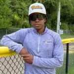 Jared Bell leaning against a yellow fence, wearing a cap and sunglasses, with greenery in the background.