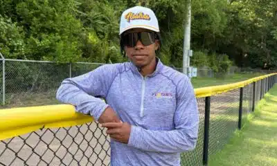 Jared Bell leaning against a yellow fence, wearing a cap and sunglasses, with greenery in the background.