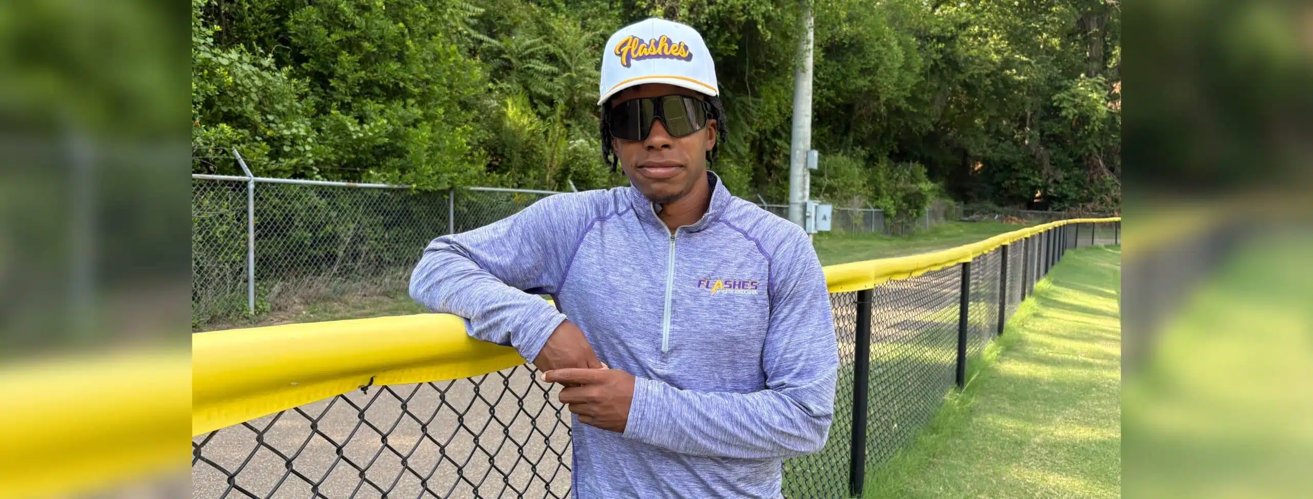Jared Bell leaning against a yellow fence, wearing a cap and sunglasses, with greenery in the background.