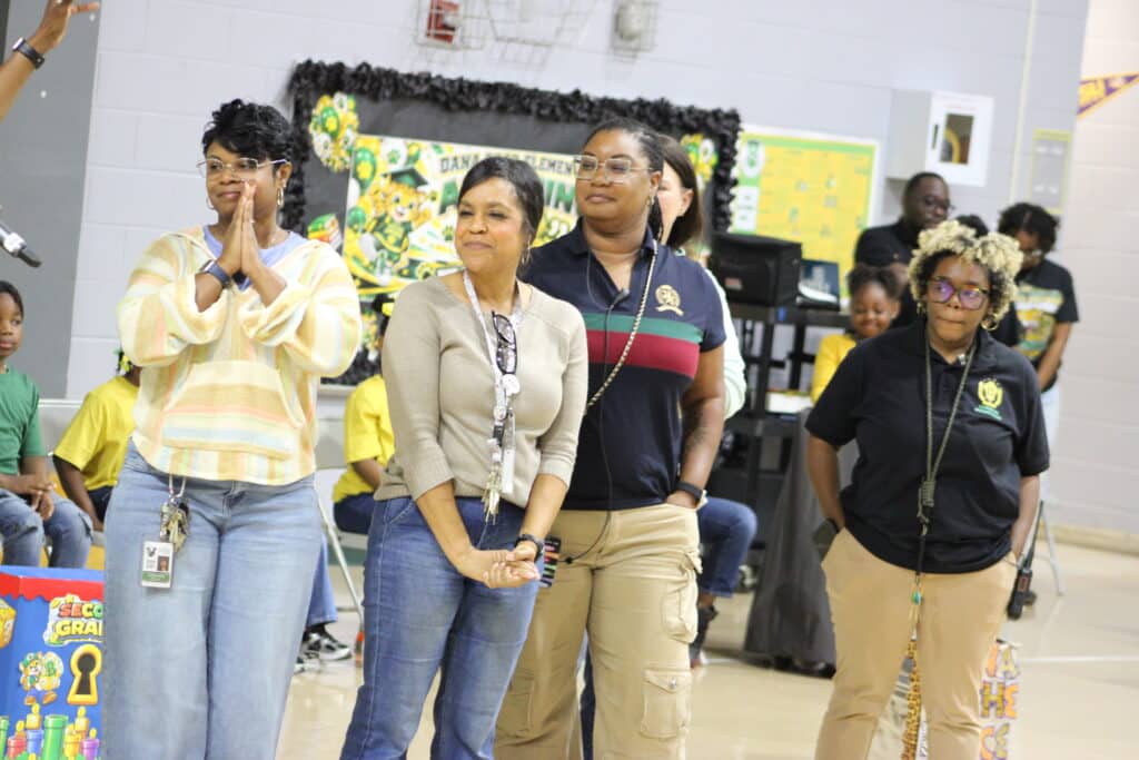 Dana Road teachers and staff stand in a school gym as one woman applauds and students sit nearby.