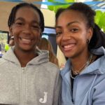 Teen with a genetic disorder smiles beside a woman at an indoor gathering with green and blue balloons.