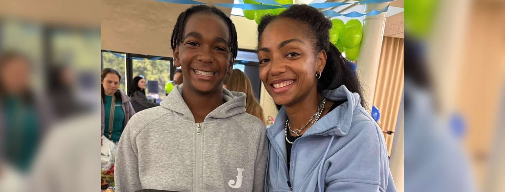 Teen with a genetic disorder smiles beside a woman at an indoor gathering with green and blue balloons.