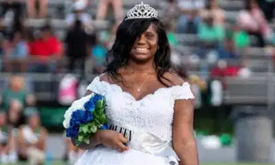 Jenieve Moorehead wearing a crown and white dress, smiling while holding a bouquet at a football game.