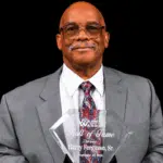 Harry Ferguson Sr. holding his Hall of Fame award, dressed in a gray suit with a patterned tie.