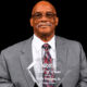 Harry Ferguson Sr. holding his Hall of Fame award, dressed in a gray suit with a patterned tie.