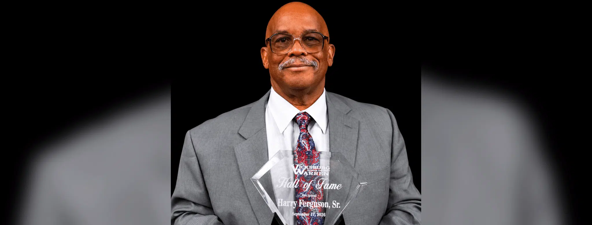 Harry Ferguson Sr. holding his Hall of Fame award, dressed in a gray suit with a patterned tie.