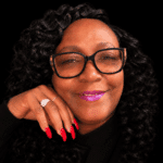 A woman with curly hair and glasses smiles, showcasing her long red nails and a ring. — Stephanie Cherey Jones