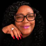 A woman with curly hair and glasses smiles, showcasing her long red nails and a ring. — Stephanie Cherey Jones