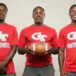 Three coaches in red shirts holding a football, standing together in a sports set — Devonte Cage and Laurence Sullivan Jr.