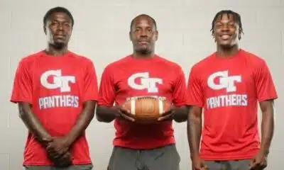 Three coaches in red shirts holding a football, standing together in a sports set — Devonte Cage and Laurence Sullivan Jr.