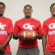 Three coaches in red shirts holding a football, standing together in a sports set — Devonte Cage and Laurence Sullivan Jr.