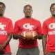 Three coaches in red shirts holding a football, standing together in a sports set — Devonte Cage and Laurence Sullivan Jr.