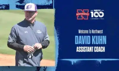 David Kuhn in a gray jacket and baseball cap, smiling on a softball field.
