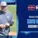 David Kuhn in a gray jacket and baseball cap, smiling on a softball field.