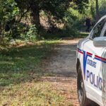 Police car with lights on near a wooded area where a person is seated on the ground, indicating a shoo — Shot in Vicksburg