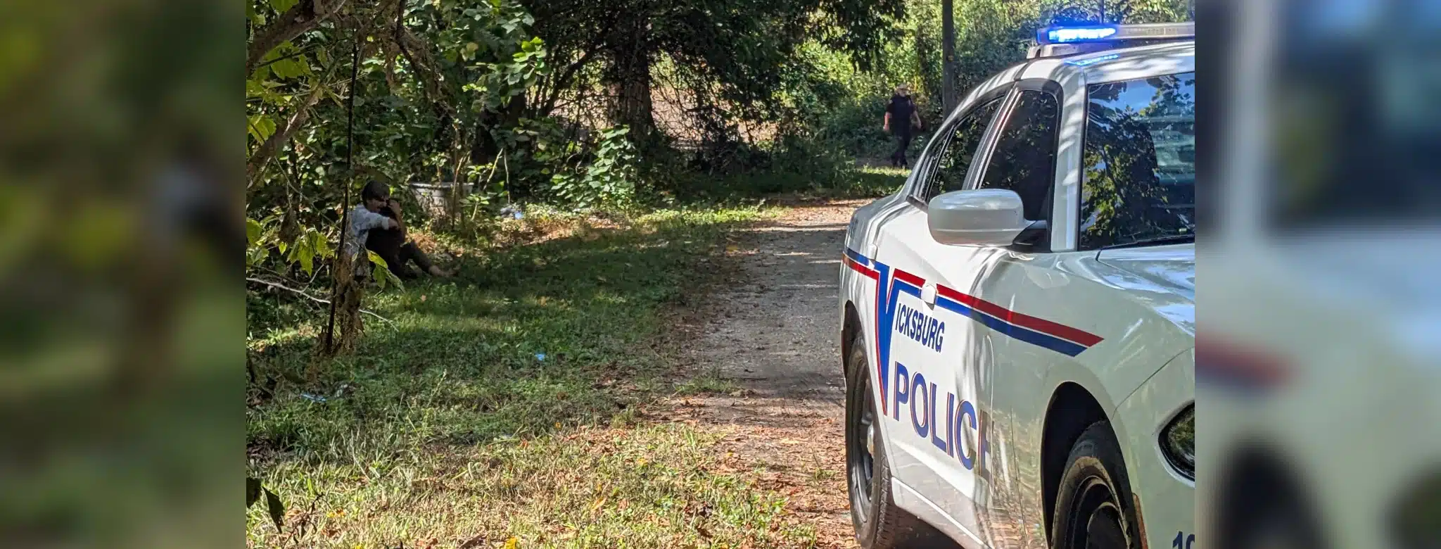 Police car with lights on near a wooded area where a person is seated on the ground, indicating a shoo — Shot in Vicksburg
