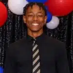 A smiling young man in a black shirt and tie stands in front of red, white, and blue balloons. — Henry Trisby III