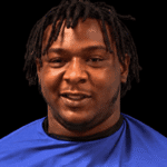 A 24-year-old man with dreadlocks wearing a blue shirt, smiling against a dark background. — 24 year old