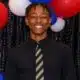 A smiling young man in a black shirt and tie stands in front of red, white, and blue balloons. — Henry Trisby III
