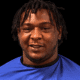 A 24-year-old man with dreadlocks wearing a blue shirt, smiling against a dark background. — 24 year old