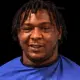A 24-year-old man with dreadlocks wearing a blue shirt, smiling against a dark background. — 24 year old