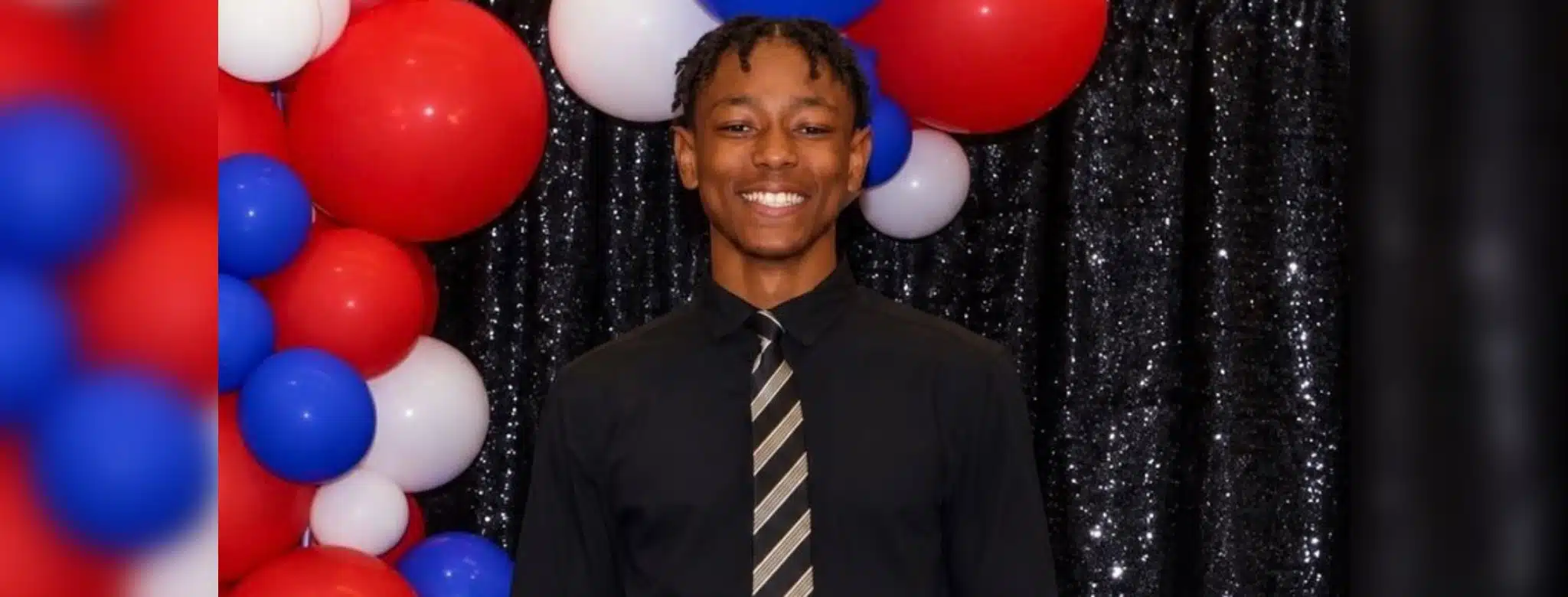 A smiling young man in a black shirt and tie stands in front of red, white, and blue balloons. — Henry Trisby III