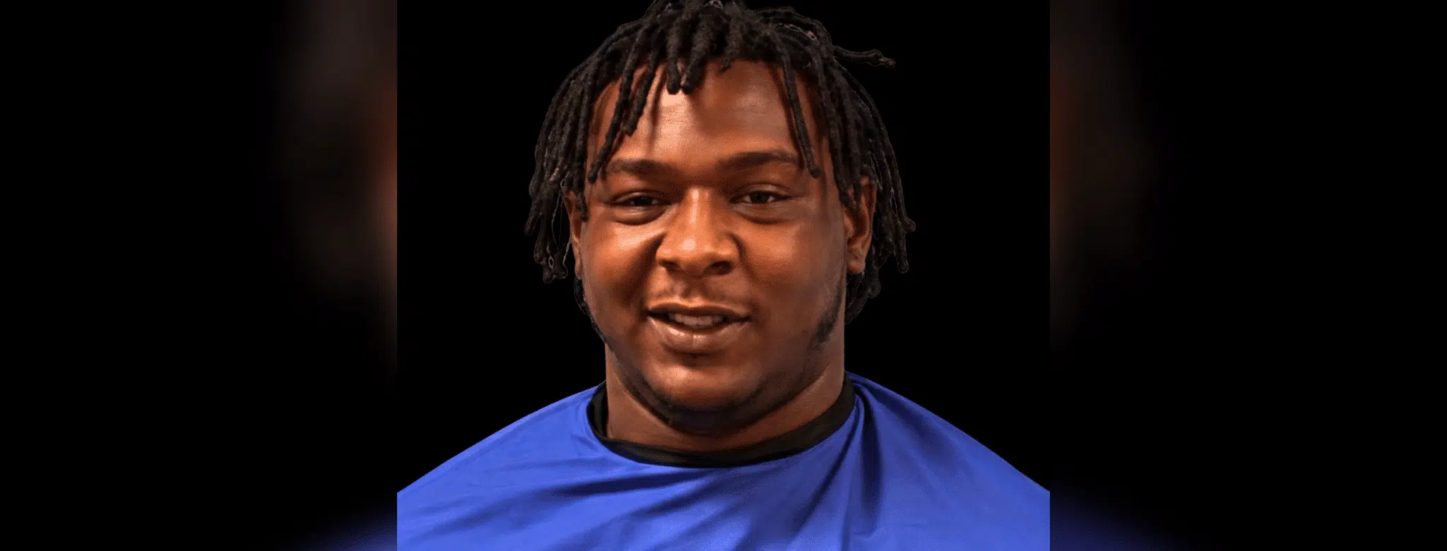 A 24-year-old man with dreadlocks wearing a blue shirt, smiling against a dark background. — 24 year old