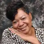 Smiling woman with short hair and a leopard print top, resting her chin on her hand against a textured background. — Verna