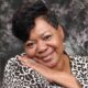 Smiling woman with short hair and a leopard print top, resting her chin on her hand against a textured background. — Verna