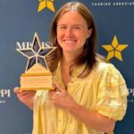 A woman in a yellow blouse holds a star-shaped award in front of a blue backdrop with Mississippi Tourism Associ — Anchuca