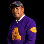 Ernest Young wearing a purple and gold sweater and dark cap against a black background