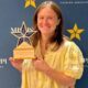 A woman in a yellow blouse holds a star-shaped award in front of a blue backdrop with Mississippi Tourism Associ — Anchuca