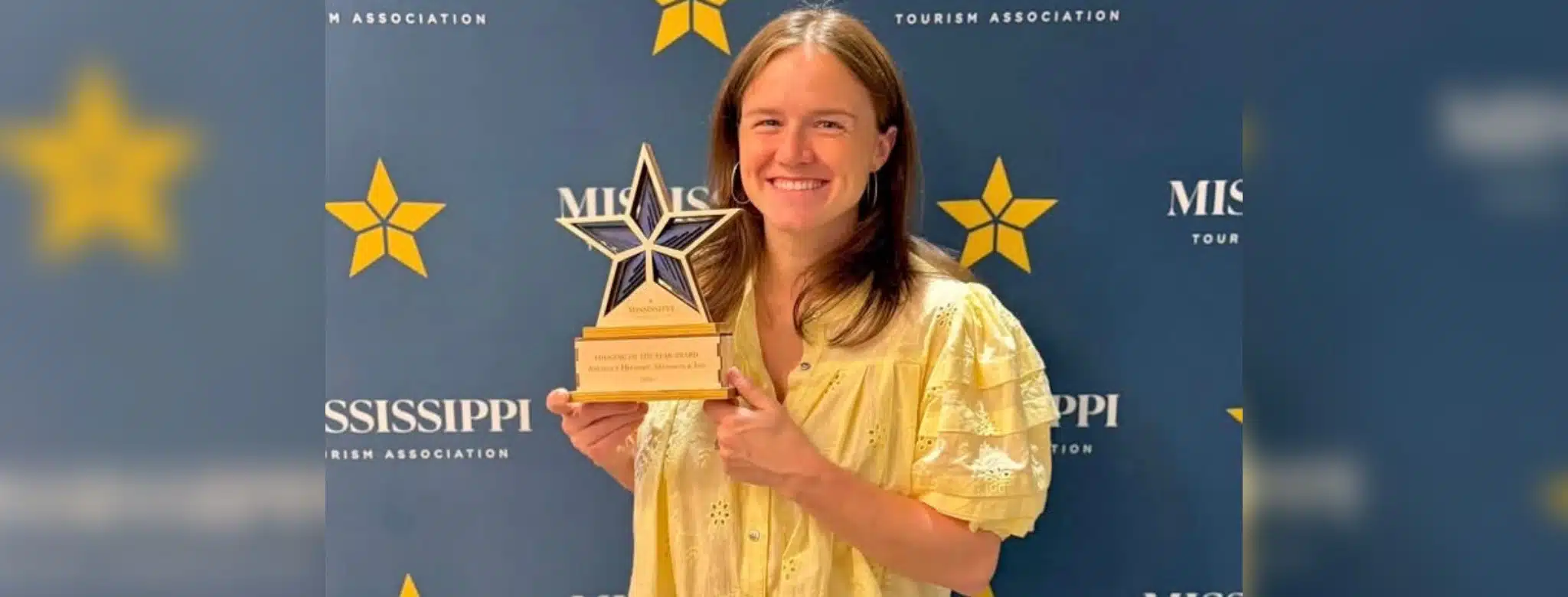 A woman in a yellow blouse holds a star-shaped award in front of a blue backdrop with Mississippi Tourism Associ — Anchuca