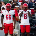 Four football players in red and white uniforms stand on the sidelines during a game. — Warren Central district play