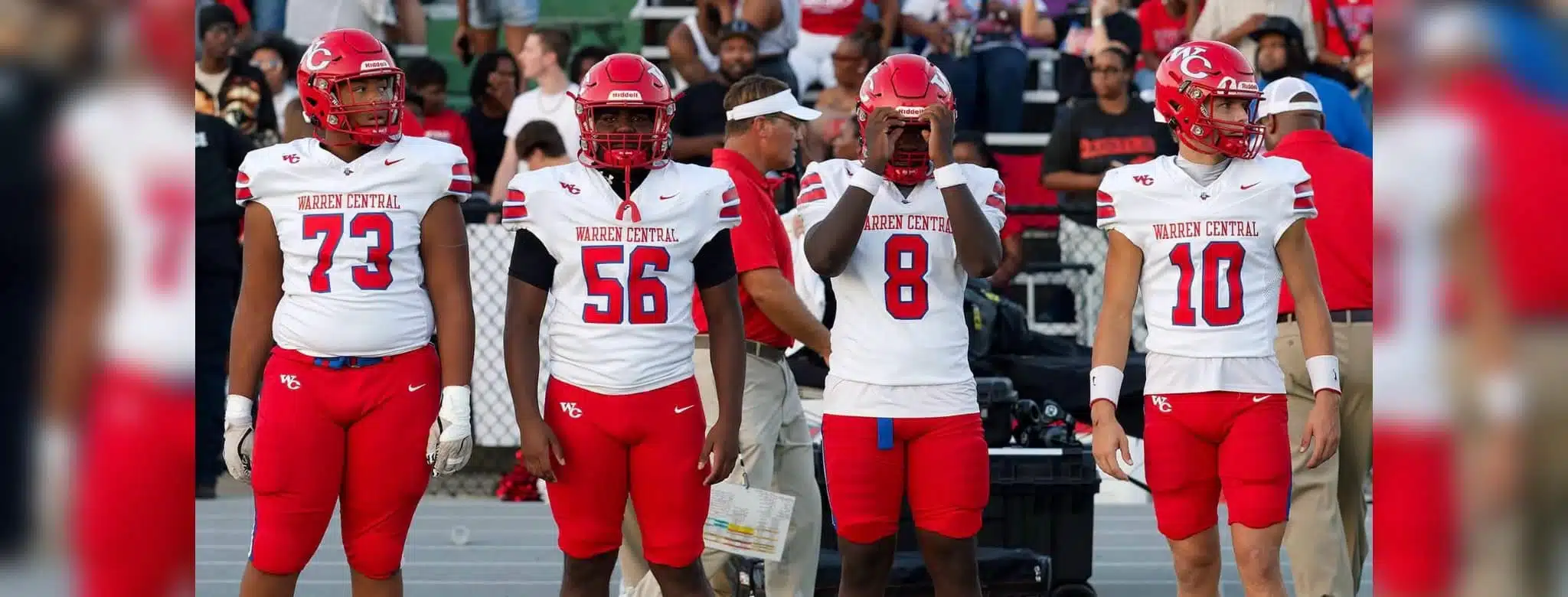 Four football players in red and white uniforms stand on the sidelines during a game. — Warren Central district play