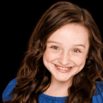 Elvie Grace smiles in a blue top in a studio portrait with a dark background.