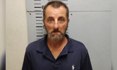 A man with a beard and blue polo shirt stands against a height chart in a police booking photo. — Shooting girlfriend